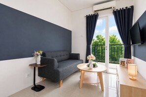 Ein modernes Wohnzimmer mit grauem Sofa, einem kleinen runden Tisch, einem an der Wand montierten Fernseher und einem Balkon mit Vorhängen.