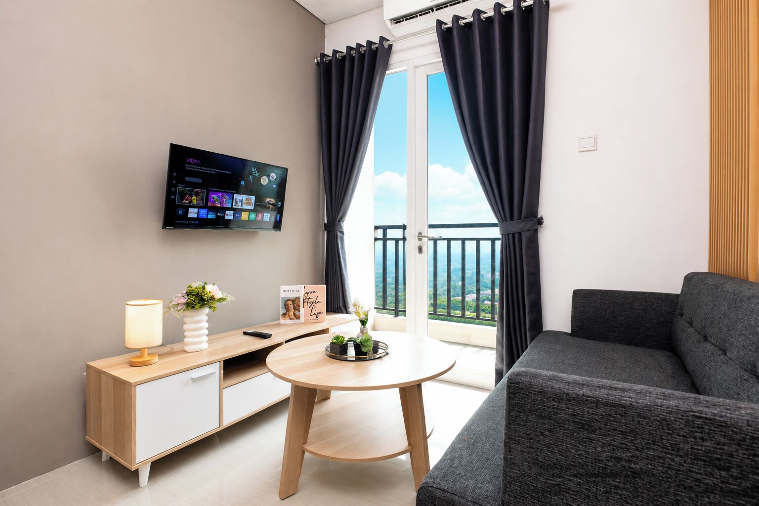 A modern living room with a sofa, a small round table, a TV mounted on the wall, and a balcony with curtains.