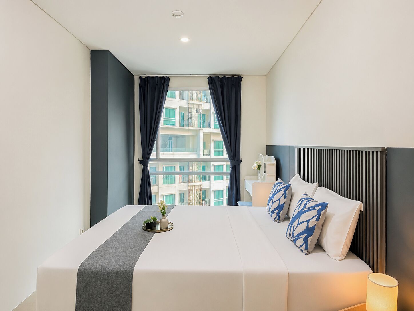 A hotel room with a bed, a bedside table, a lamp, and a view of a building through the window.