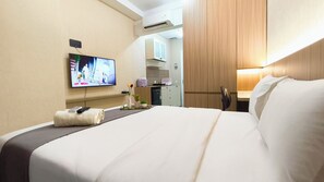 A hotel room with a bed, a TV mounted on the wall, a small kitchenette, and a bedside table with a remote control and a vase with flowers. - Modern Look Studio At Apartment The Vertu (Surabaya)