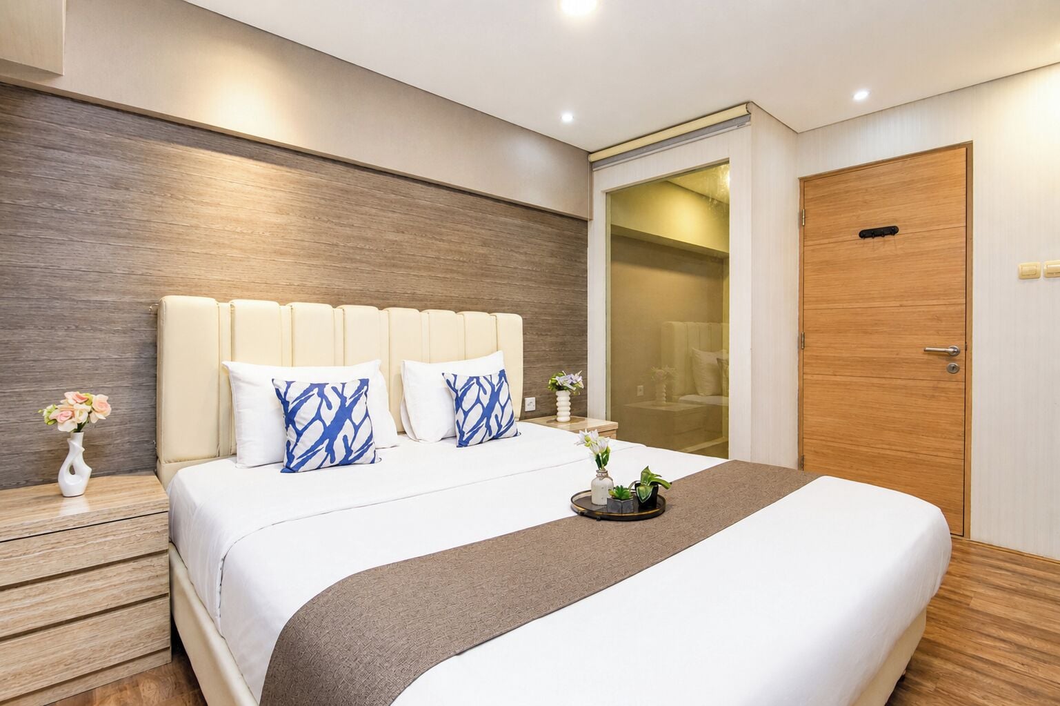 A modern hotel room with a large bed, a wooden headboard, a nightstand with a vase of flowers, and a wooden door.