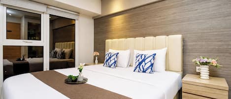 A modern hotel room with a large bed, a wooden headboard, a bedside table with a vase of flowers, and a view through a sliding door.