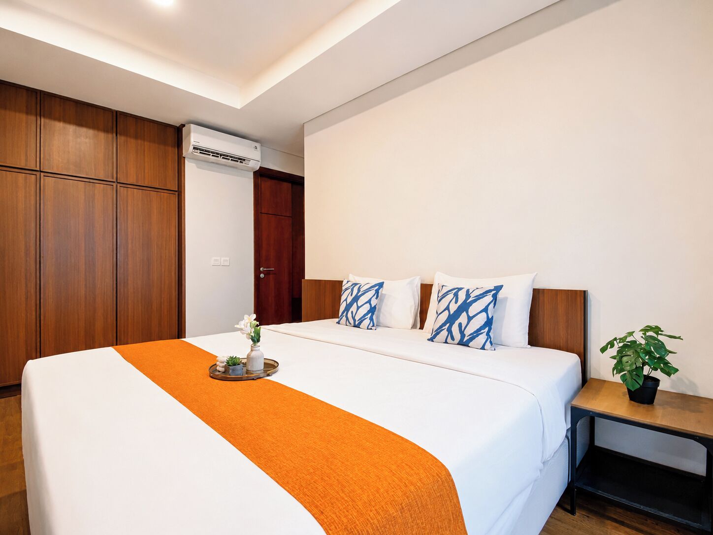 A hotel room with a bed, a bedside table with a plant, and a wooden wardrobe.