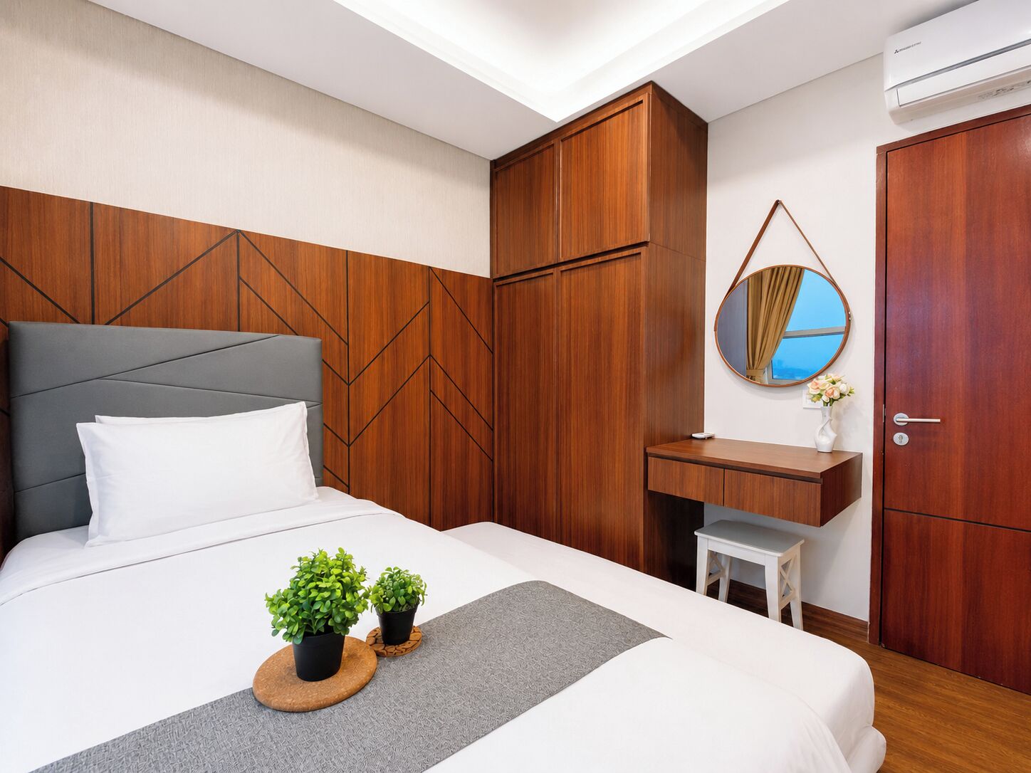 A hotel room with a bed, wooden wardrobe, bedside table, mirror, and an air conditioning unit.
