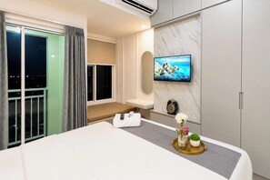 Ein Hotelzimmer mit Bett, einem an der Wand montierten Fernseher, einem Nachttisch mit Fernbedienung und Blumen, sowie Blick auf die beleuchtete Stadt durch das Fenster.