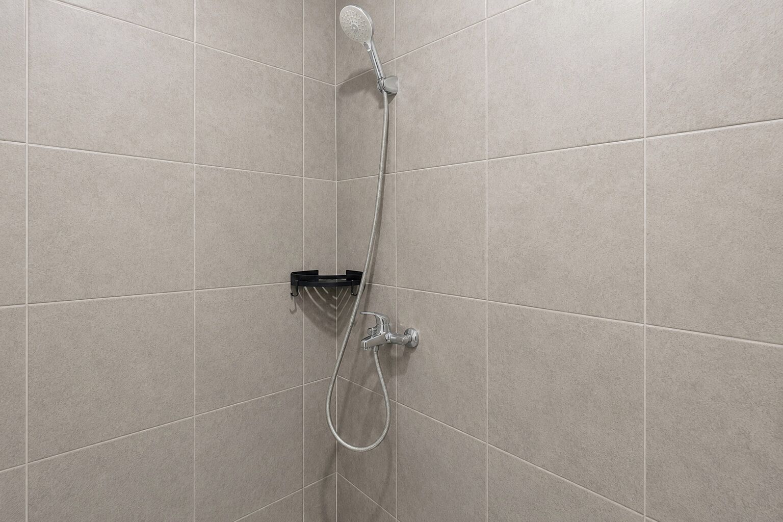 A shower with a handheld showerhead and a wall-mounted faucet against a tiled wall.