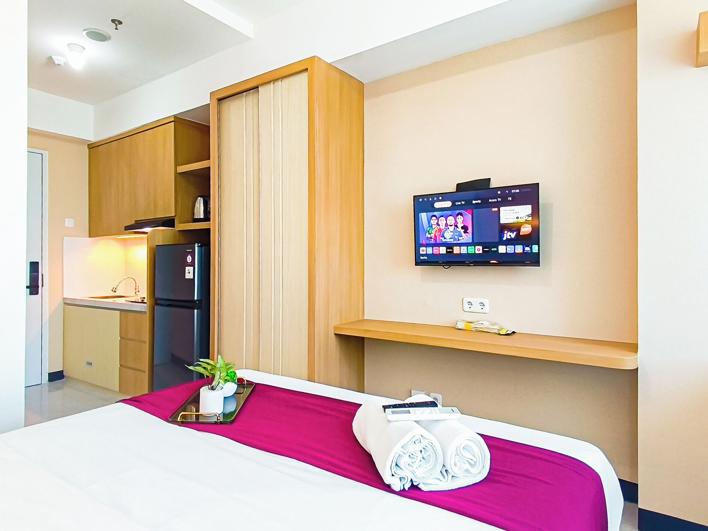 A hotel room with a bed, a TV mounted on the wall, a small shelf, and a kitchenette area.