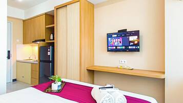 A hotel room with a bed, a TV mounted on the wall, a small shelf, and a kitchenette area.