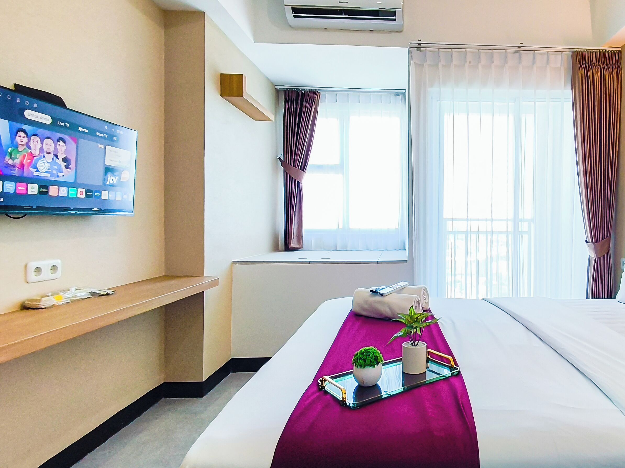 A hotel room with a bed, a TV on the wall, a window with curtains, and a small shelf with items.