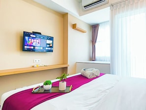 A hotel room with a bed, a mounted TV displaying a sports channel, a small plant, and a tray with a remote control. - Well Appointed Studio At Apartment Westown View Surabaya (Surabaya)