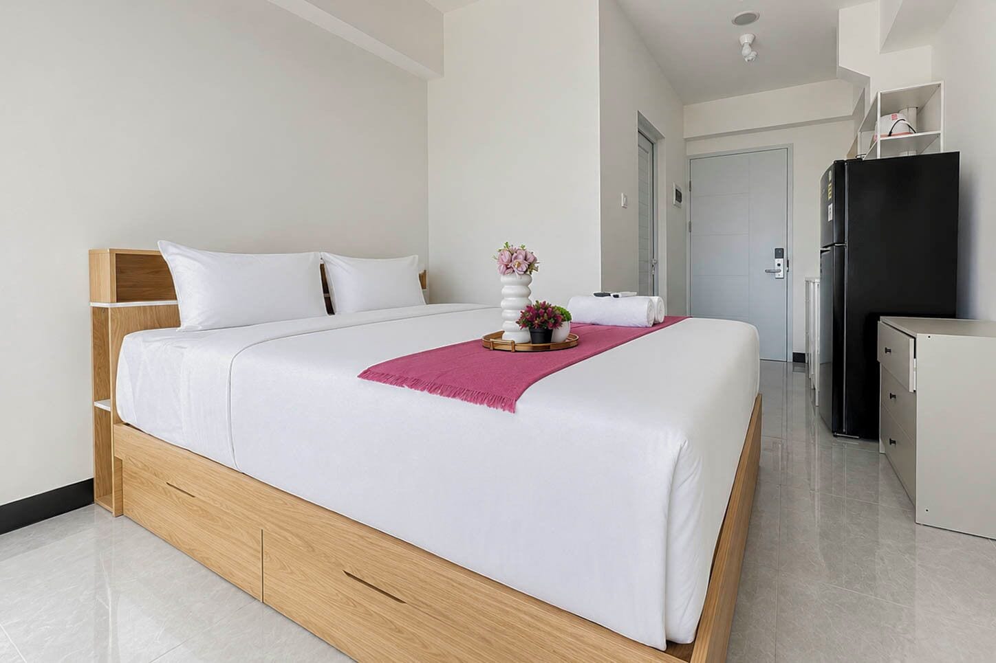 Ett modernt hotellrum med en stor säng, ett träfärgat sänggavel, ett nattduksbord med en vas med blommor och ett svart kylskåp.