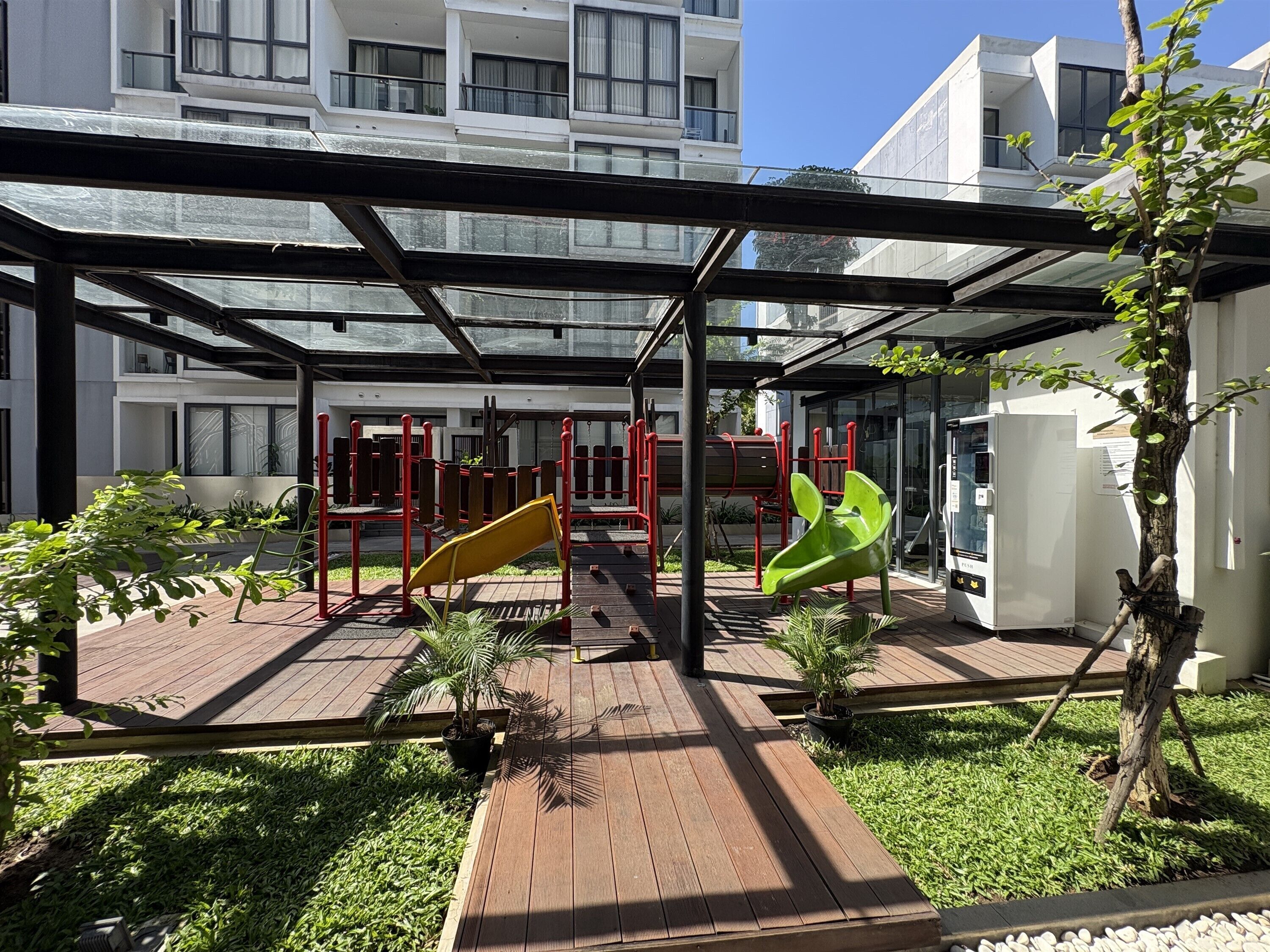 Un espacio exterior techado con un parque infantil, un refrigerador y un árbol.