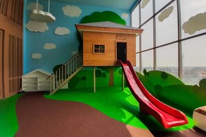 A children's play area with a slide, a wooden playhouse, and a view through large windows. - Bright Comfortable Studio Room At Apartment Springwood Residence Tangerang (Tangerang)