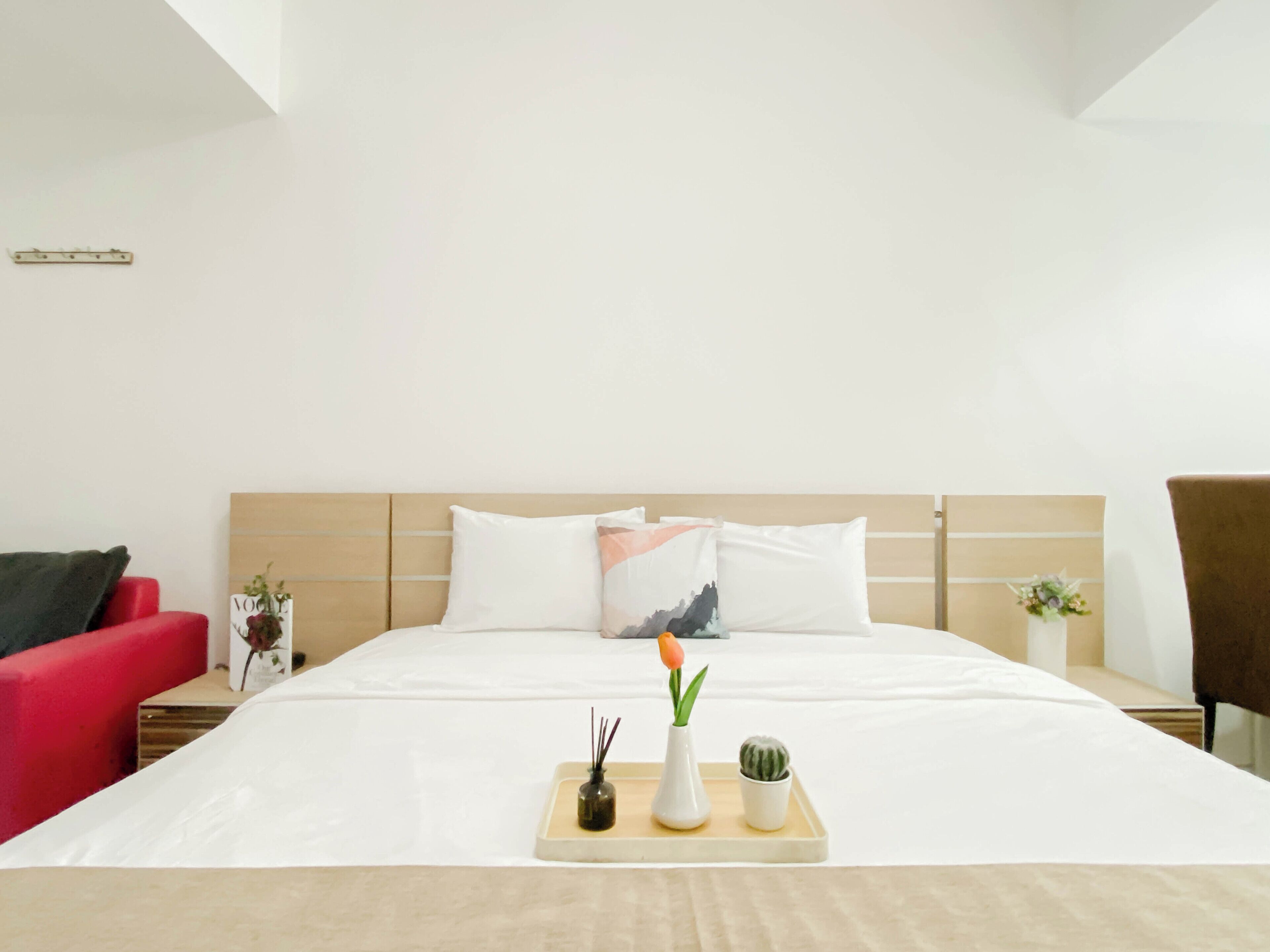 A hotel room with a large bed, a red chair, a nightstand with a vase, and a small table with a vase and a plant.