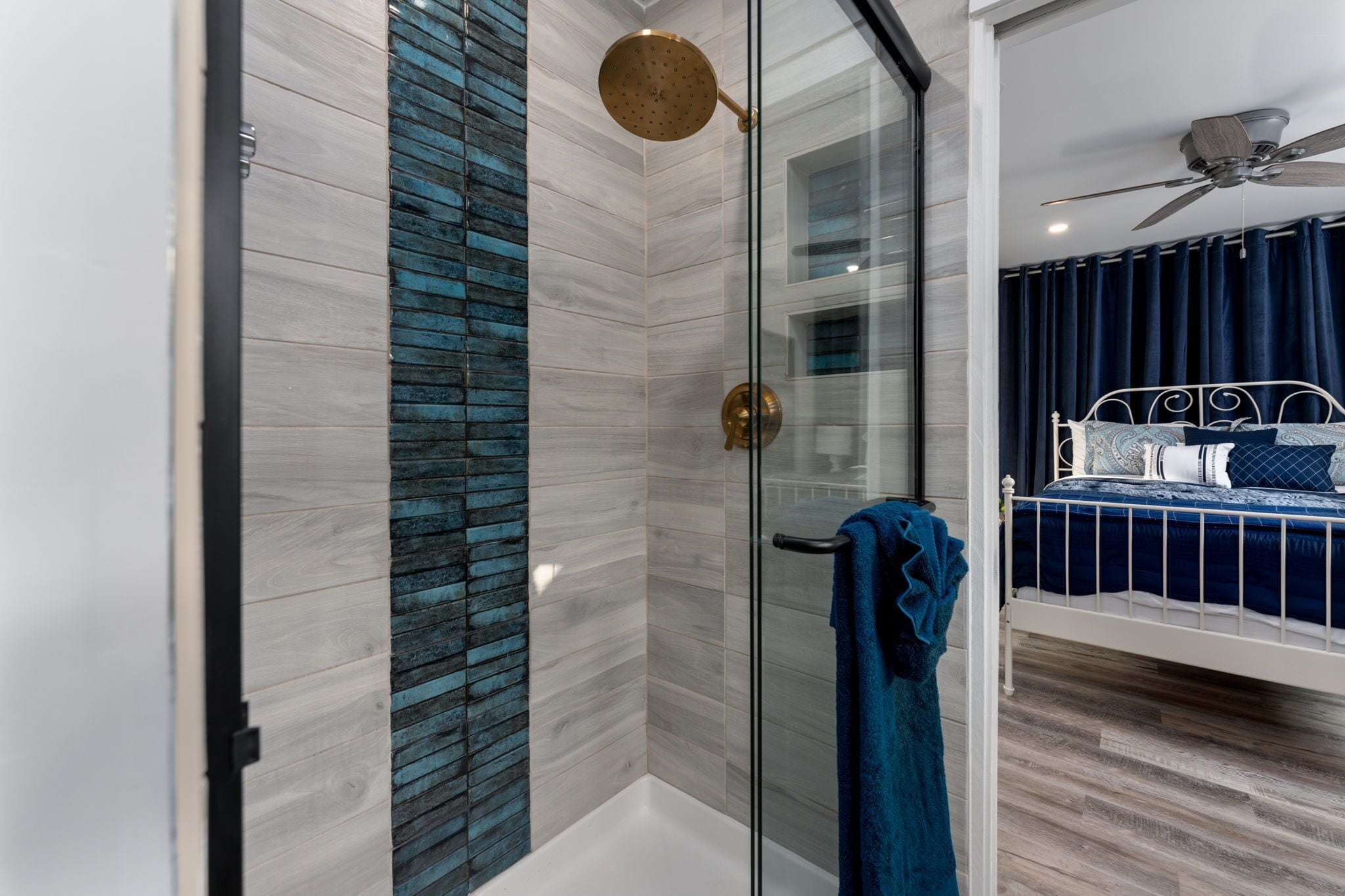 A modern bathroom with a glass shower enclosure, a white bathtub, and a bed with a blue headboard.