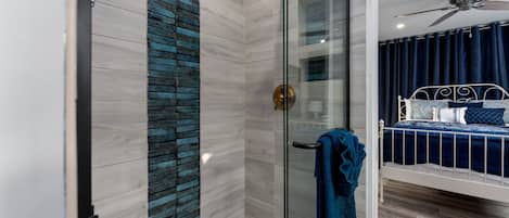 A modern bathroom with a glass shower enclosure, a white bathtub, and a bed with a blue headboard.