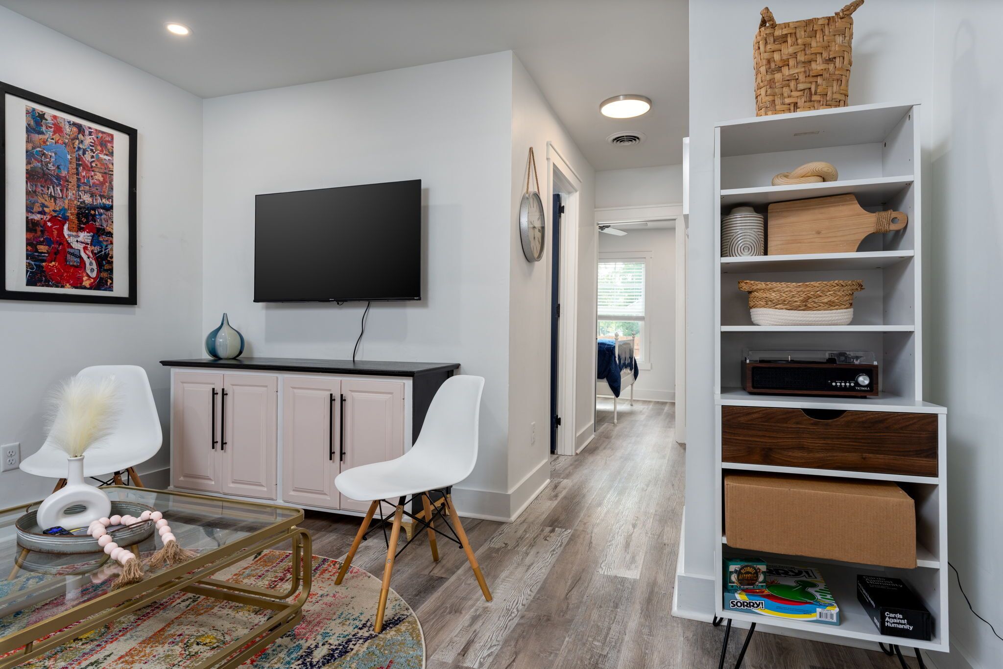 Uma sala de estar moderna com televisão de tela plana, cadeiras brancas, uma mesa de centro de vidro, um vaso decorativo, um relógio e uma estante com diversos objetos.