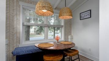 Uma área de refeições com mesa redonda, bancos, luminárias pendentes trançadas e uma janela com persianas.