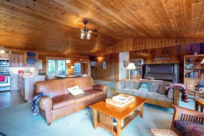 Ein gemütliches Blockhäuschen-Interieur mit Holzbalkendecke, einem Wohnzimmer mit Sofa und Couchtisch, einer Küchenzeile und einem Fernseher.