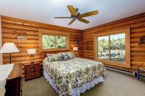 Ein Schlafzimmer in einer Blockhütte mit einem Bett, zwei Fenstern, einem Deckenventilator und einem Nachttisch mit Lampen.