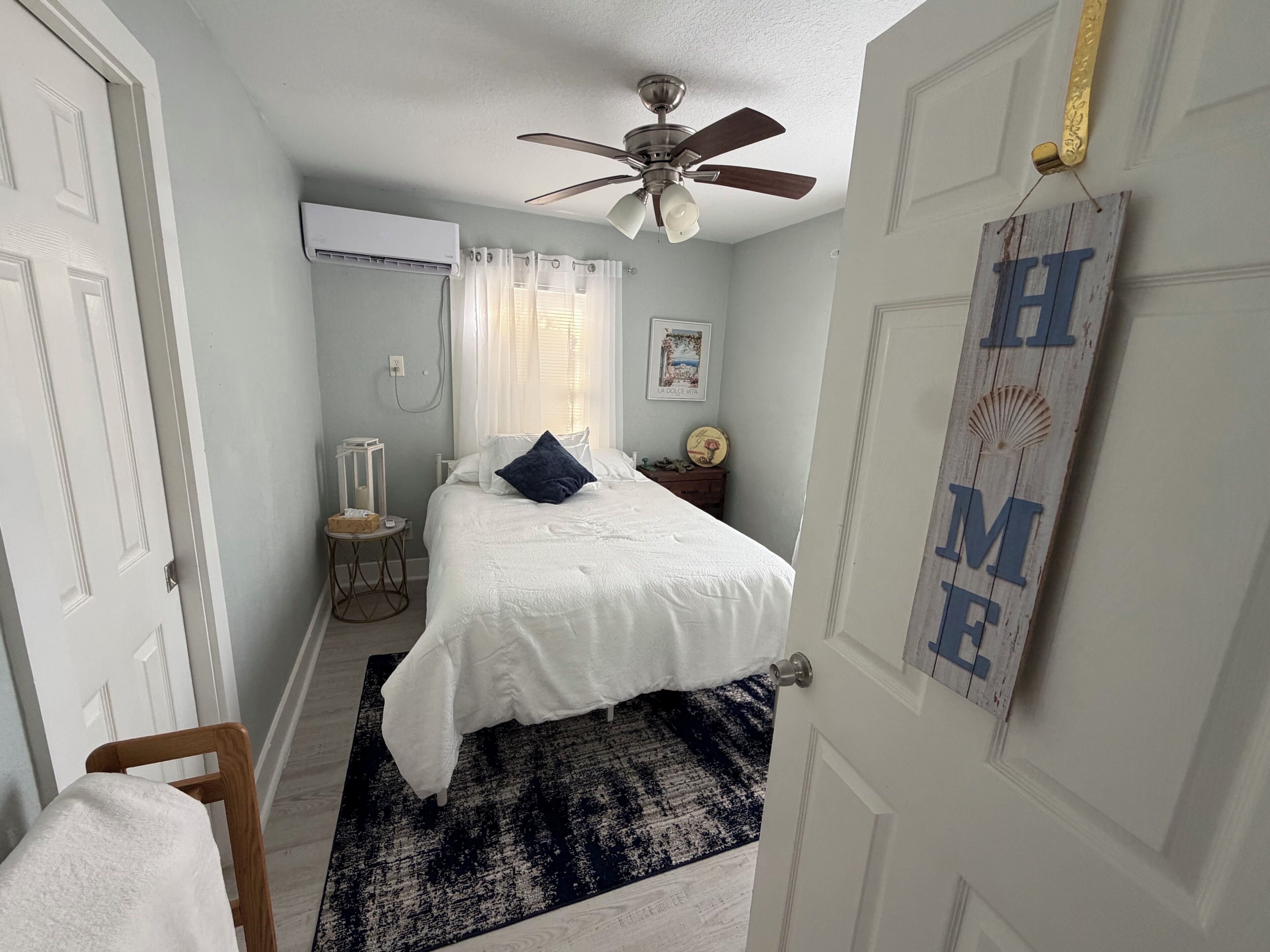 A bedroom with a bed, a ceiling fan, a window with curtains, a bedside table, and a wall-mounted sign.