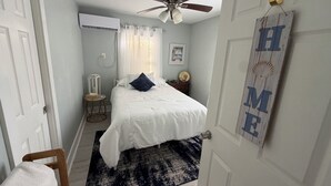 Um quarto com cama, ventoinha no teto, janela com cortinas, mesa de cabeceira e um aviso fixado na parede.
