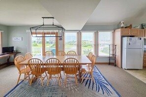 A dining area with a table and chairs, a kitchen with a refrigerator, and large windows with blinds. - 7853 - Anchor Management Spacious 6BR, 5.2BA oceanside home in Frisco for up to 12 guests. (Frisco)