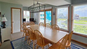 A dining area with a rectangular table and six chairs, a chandelier, and large windows with blinds. - 7853 - Anchor Management Spacious 6BR, 5.2BA oceanside home in Frisco for up to 12 guests. (Frisco)