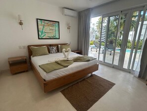 Un dormitorio con cama, mesita de noche, aire acondicionado instalado en la pared, un cuadro y vistas a una piscina y palmeras a través de un ventanal corredero.