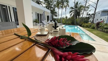 Uma mesa de madeira com um jogo americano, uma chaleira branca, duas xícaras brancas e uma flor vermelha.