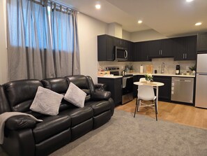 A modern living room with a black leather sofa and a small white table, a kitchen with dark cabinets and stainless steel appliances, and a window with grey curtains. - PRIVATE 2 Bedroom Basement Suite | Near Foothills | Airport | Banff & Attraction (Calgary)