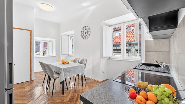 A modern kitchen with a central island, a dining area with a table and chairs, and a view of a red-tiled roof through a window. - Apartment Gracija  2-Bedroom Apartment in Dubrovnik Old Town
 (Dubrovnik)