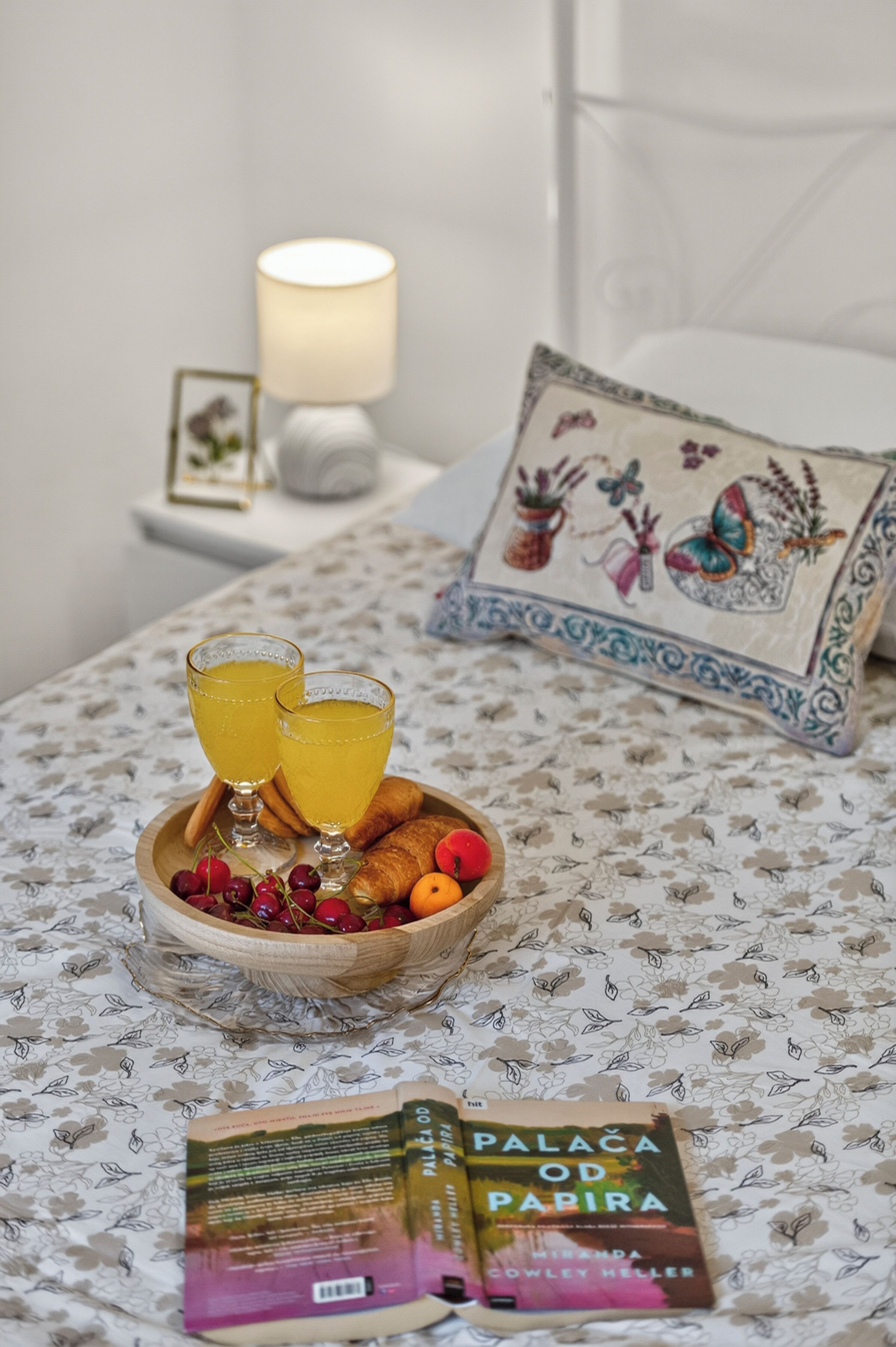 A bed with a floral-patterned bedspread, a wooden tray with fruit and two glasses of orange juice, a book titled 'Palaça do Papira,' and a decorative pillow with a floral design.