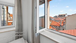 A room with a window overlooking a cityscape, a bed, and a curtain. - Apartment Gracija  2-Bedroom Apartment in Dubrovnik Old Town
 (Dubrovnik)