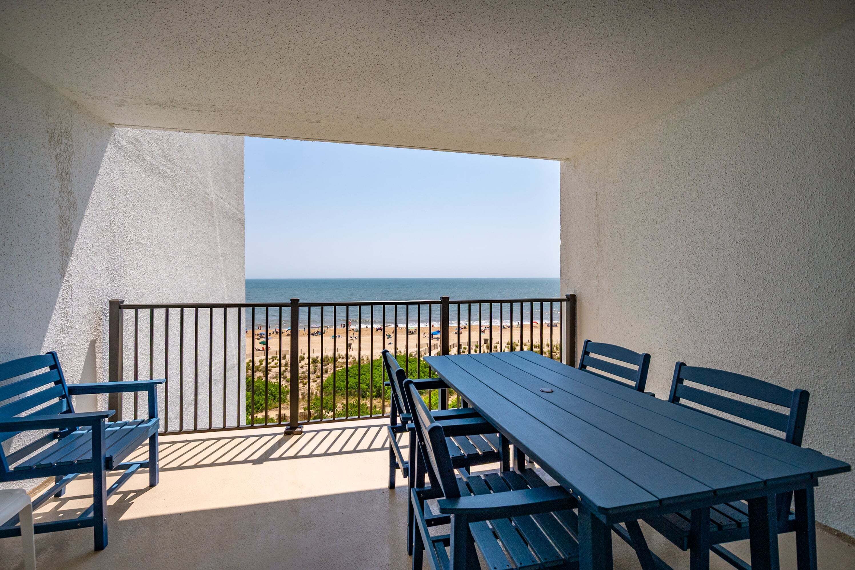 A balcony with a table and chairs, a view of the beach, and a clear sky.