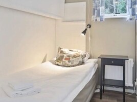 Una habitación pequeña con una cama individual, colchón blanco, almohada con diseño de periódico, mesita de noche con lámpara y ventana con cortinas.