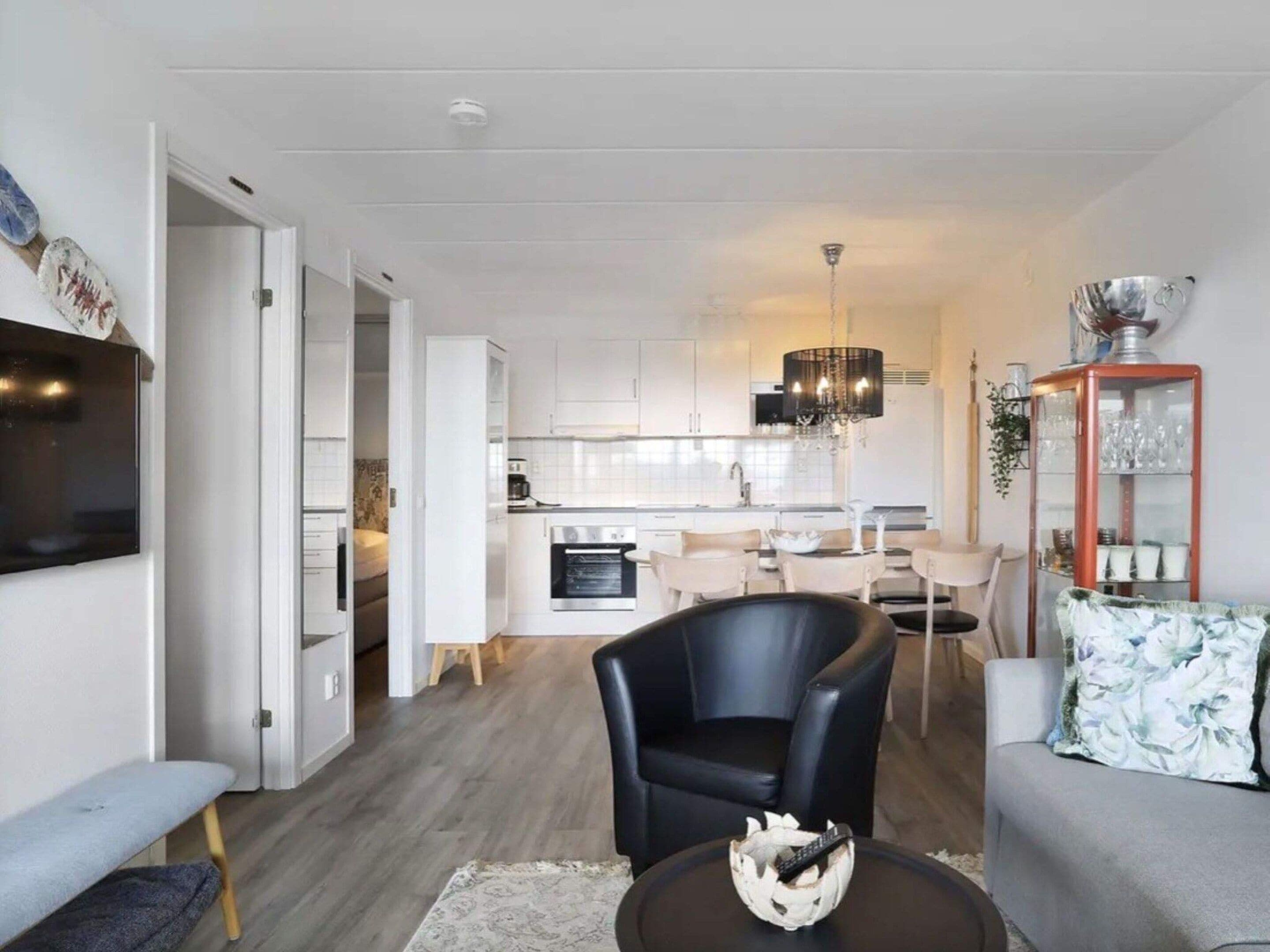 A modern living room with a sofa, a coffee table, and a TV. There is a dining area with chairs and a chandelier. The kitchen is visible in the background with white cabinets and a stove.