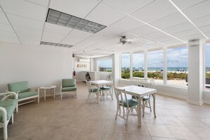Ein geräumiger Raum mit großem Fenster und Blick zum Strand, weiße Decke mit Lüftungsgitter sowie ein weißer Tisch mit Stühlen.