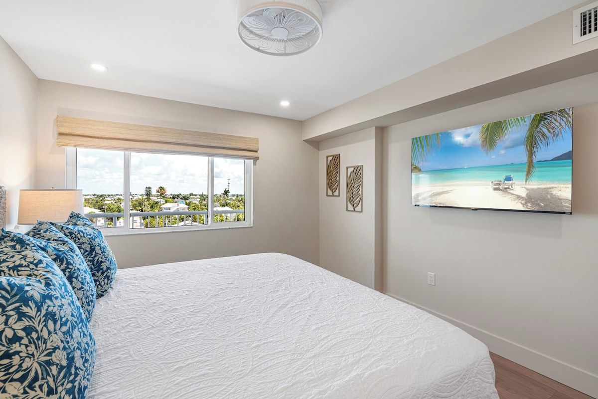 A bedroom with a large bed, a window with a view of a beach, and a painting of a beach scene.