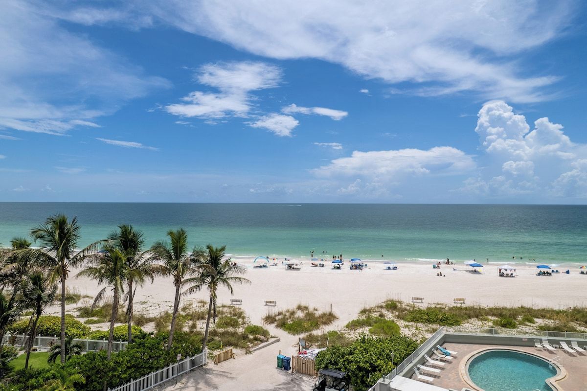 A beach view with a pool area, palm trees, and a clear sky.