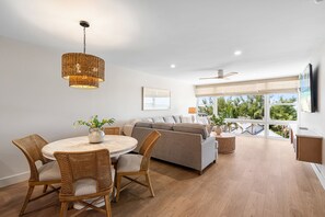 Ein modernes Wohnzimmer mit einem runden Esstisch, Korbstühlen, einem grauen Sofa und einem großen Fenster mit Blick.