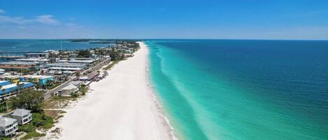 An aerial view of a beach with a clear blue sky, turquoise waters, and a row of buildings along the shore.