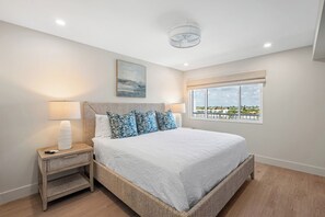 A bedroom with a bed, two bedside tables, a ceiling fan, a window with a view, and a painting on the wall. - Martinique South 506 (Holmes Beach)