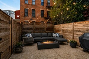 En terrasse med en sort sofa, et bålsted og lyskæder.