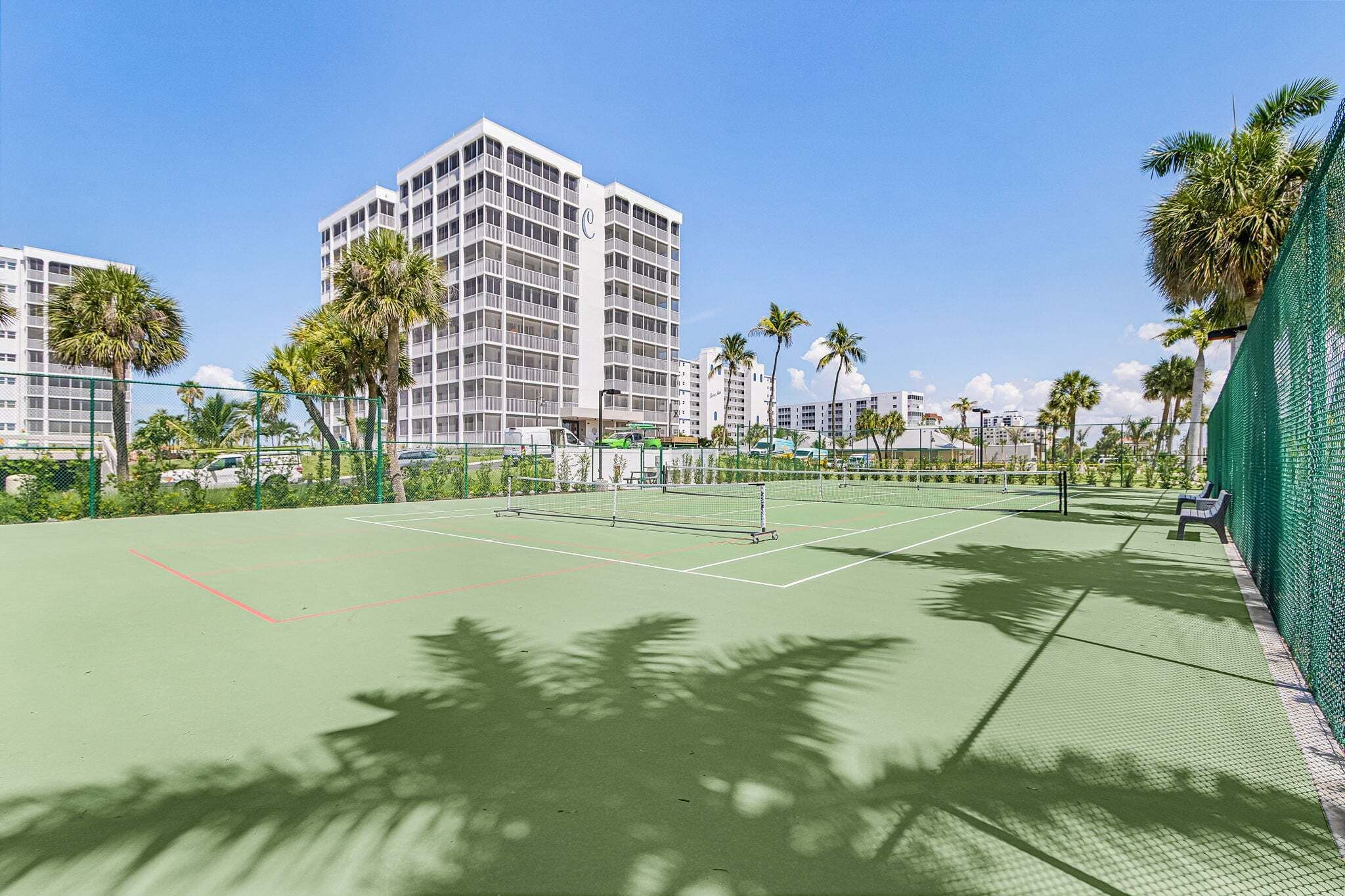 A tennis court with a net, surrounded by palm trees and a building in the background.