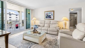 Un soggiorno moderno con divano, poltrona, tavolino in vetro e due lampade da terra. Un quadro è appeso al muro, e si intravede un balcone con tavolo e sedie.