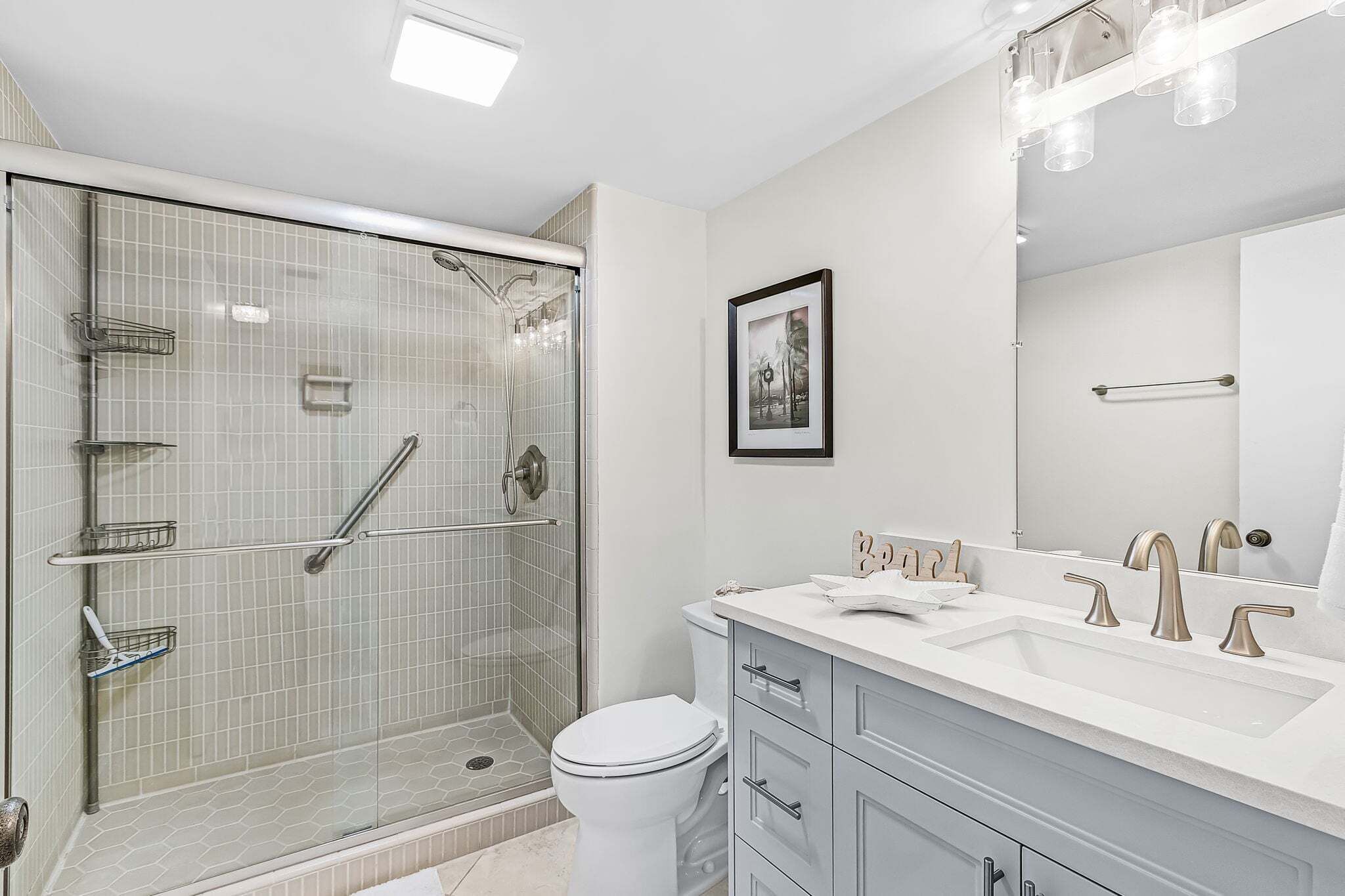 A modern bathroom with a glass-enclosed shower, a white toilet, a vanity with a mirror, and a framed picture on the wall.