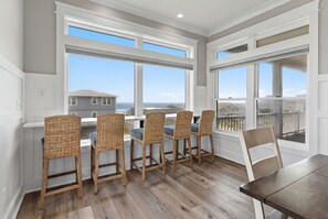 Un comedor con cuatro sillas de mimbre, una mesa de madera y ventanas amplias con vistas al océano y casas vecinas.