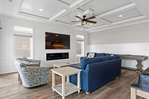 A living room with a blue sofa, a patterned armchair, a small table, a ceiling fan, and a fireplace. - Weekend Rescue: 10BR 10.5BA, Ocean Views, Pool, Hot Tub, Elevator, Dog Friendly, Sleeps 22 (Oak Island)
