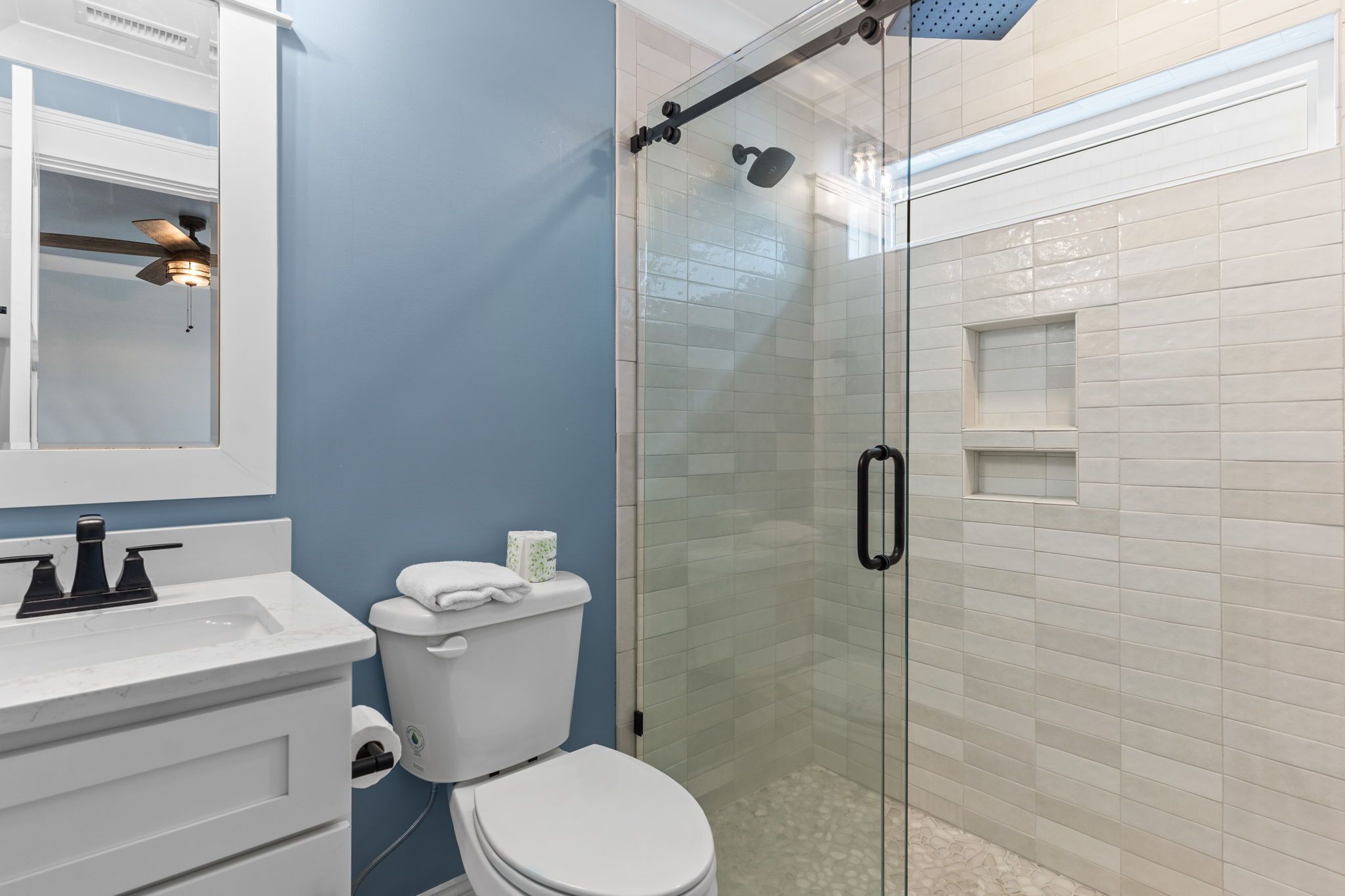 A bathroom with a glass-enclosed shower, a white toilet, a sink with a mirror, and a ceiling fan.